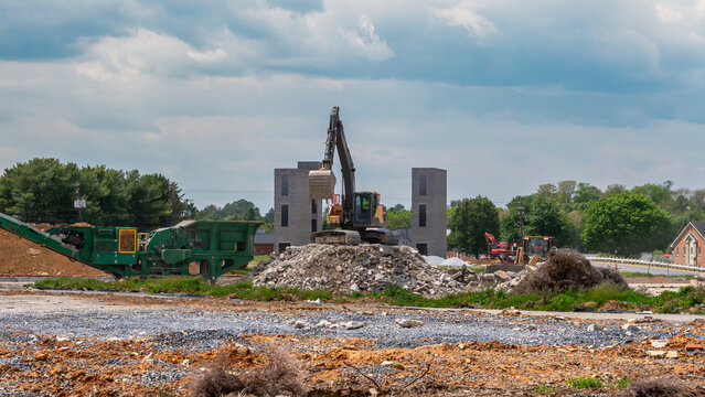 Heavy machinery is working at a construction site in a rural area. Large rocks and dirt are moved around. New structures are being built among green trees and cloudy skies.