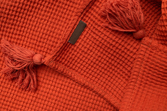 Red and orange throw blanket with decorative tassels and blank logo label close-up. Cotton blanket mockup for home decor with blank dark fabric tag. Waffle texture, cozy fabric detail.