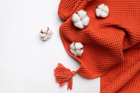 Bright red orange waffle-textured fabric with decorative tassel featuring cotton flowers on gray background top view. Natural cotton textile concept in cozy autumnal shades. Throw or towel for interio