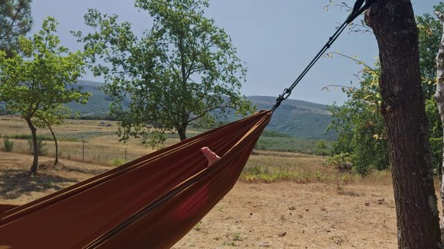 Hammock relaxation in the mountains surrounded by nature
Close view of feet gently moving while swinging in a hammock outdoors, surrounded by green trees and mountain scenery. Peaceful relaxation 