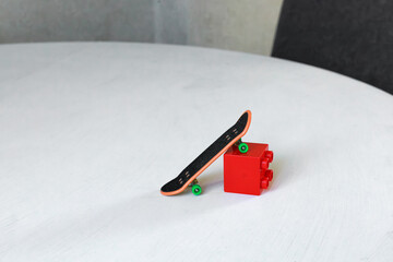 Miniature fingerboard skateboard rests on a small red block ramp on a white tabletop. Concept of fingerboarding, obstacle tricks, skatepark simulation, creative play and indoor hobby. © Natallia