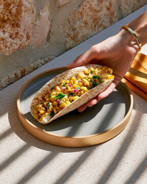 Hand holding vegetarian taco filled with grilled cheese, corn, red onion and jalapeno over plate in bright natural sunlight
