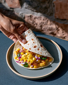 Hand preparing vegetarian taco with grilled cheese, corn, red onion and jalapeno in tortilla on plate under bright natural sunlight