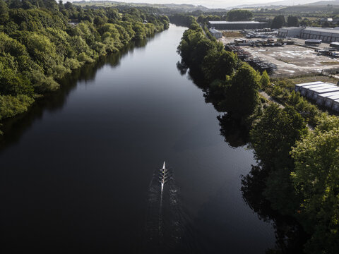 Aerial view of a rowing team in a boat on the River Lune surrounded by lush trees and industrial buildings in Lancaster, England, United Kingdom.