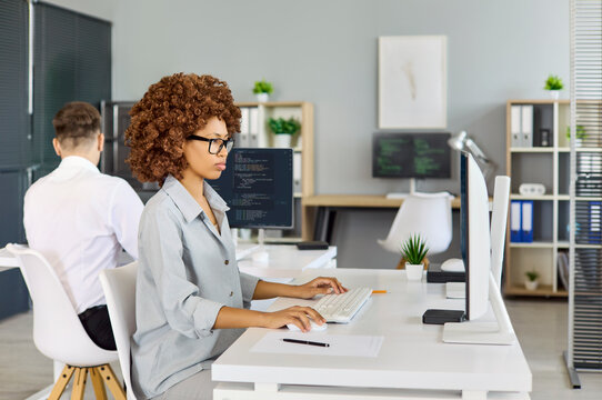 Programmer developer coding at computer in office workplace. She types at a clean desk as code fills monitors and a coworker works behind in the space. Concept of modern software development.