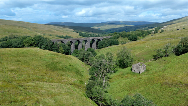Aerial view of Dent Viaduct spanning the lush green valley with rolling hills, scattered trees, and a stone structure in the foreground in Yorkshire Dales, North Yorkshire, UK.