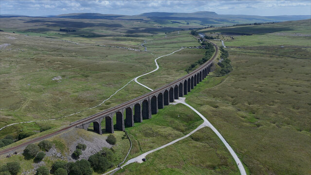 Aerial view of the historic Ribblehead Viaduct spanning the vast green landscape of the Yorkshire Dales with railway tracks and winding paths in Carnforth, England, United Kingdom.
