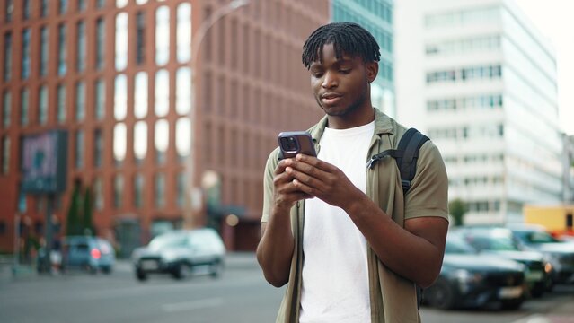 Smiling young man holding smartphone and texting message, standing outdoors in modern city street with cars and tall buildings in the background. Handsome male tapping and chatting on mobile phone.