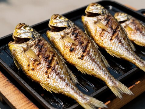 Four whole grilled yellow croaker fish sit on a black grill pan. Seafood is cooked and ready to eat with crispy skin and visible grill marks. It looks delicious and appetizing.