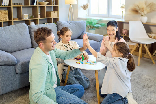 Family parents children playing boardgame together home. Smiling mother and father play with two kids at a small table in a bright livingroom, celebrating. Concept of family fun and bonding.