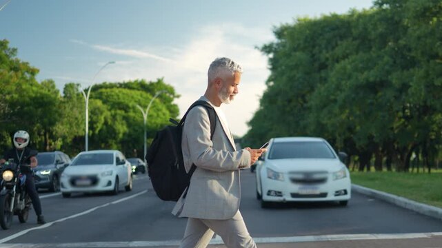 Mature Brazilian businessman with backpack using smartphone while crossing urban street on pedestrian crosswalk
