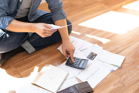 Stressed Asian man sitting on floor using calculator to check receipts and calendar for monthly expenses, financial problems, debt management, and bankruptcy concept at home.