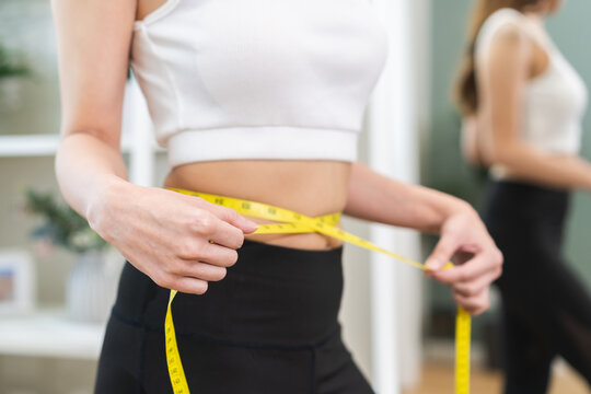 Loss weight slim body, young woman,girl measuring her waist size with tape measure, wear in fit sport, looking reflection in mirror at home. Sport strong person in gym fitness for wellbeing healthcare