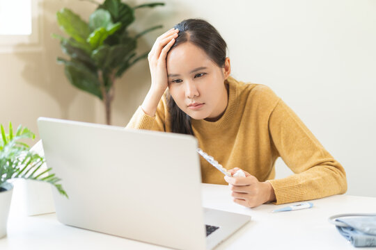 Stressed asian young woman holding a blister pack of pills and touching her head while looking at a laptop screen, consulting an online doctor about her severe headache symptoms from home.