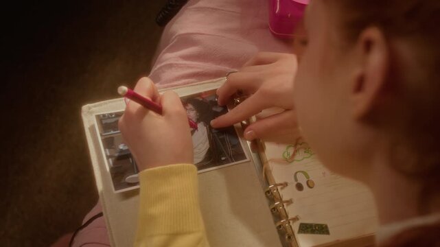 Caucasian teenage girl drawing red horns on photo of boy in her personal diary, high angle over shoulder view. Clip is perfect for youth relationship drama or mental health campaigns 