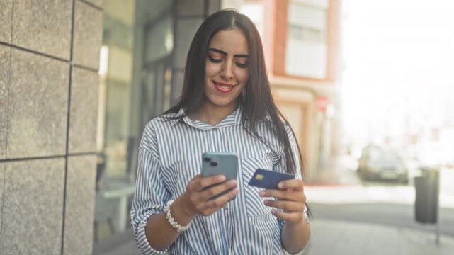 Young woman holding smartphone and creditcard while smiling on a city street beside a building; confidence convenience.