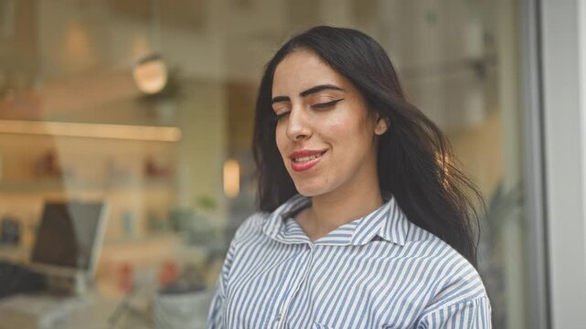 Young arab woman smiling with eyes closed and hair blowing, striped shirt and visible face at storefront building; serenity.