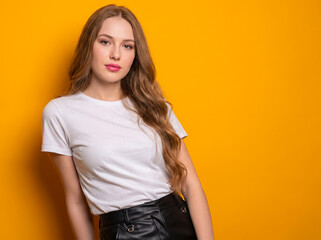 Fototapeta premium Beautiful young woman portrait in blank white t-shirt and black leather skirt posing against a bright yellow background with copy space for casual fashion mockup.