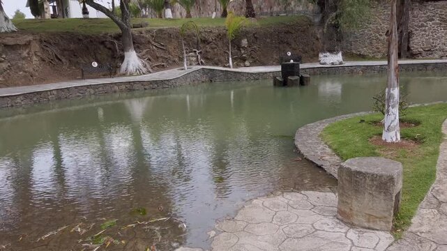 Quiet garden pond with stone walkway and green water reflection under trees in park.