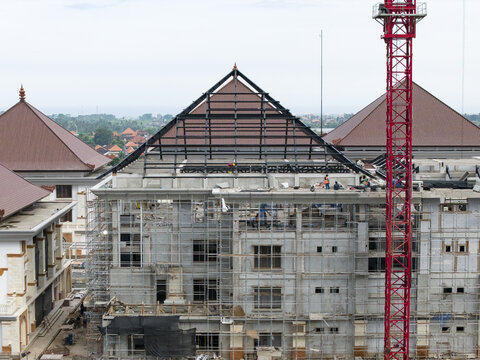Aerial view of building construction process with a prominent red crane, steel roof framework, and scaffolding surrounding the structure in Gianyar, Bali, Indonesia.