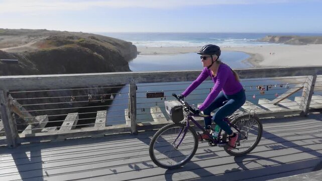 A woman rides her bike on an old train bridge in Fort Bragg, California. The ocean is visible as she passes over bridges.