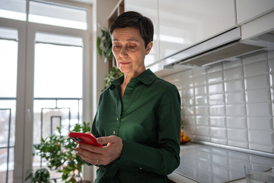 Thoughtful sad mature woman receiving bad news from smartphone message at home. Serious senior female reading SMS on mobile phone at kitchen, deep in thought, thinking, looking puzzled and worried. 