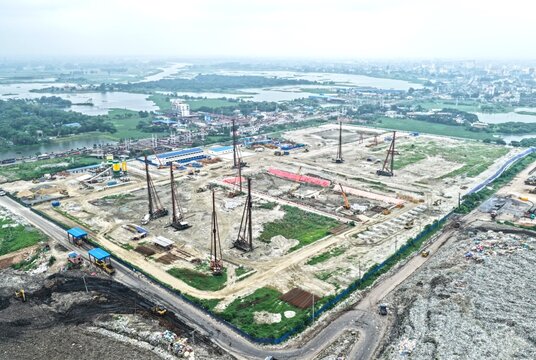 Aerial view of the Aminbazar Landfill construction site with piling rigs and heavy machinery on a large dirt plot surrounded by green fields in Aminbazar, Dhaka Division, Bangladesh.