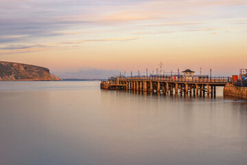 Fototapeta premium A clear November evening long exposure looking across smooth waters along Swanage Pier with a pink sky