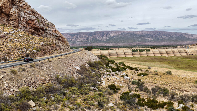 Aerial view of Little Karoo landscape featuring a winding road with a car and a large dam wall under a cloudy sky in Garden Route District Municipality, Western Cape, South Africa.