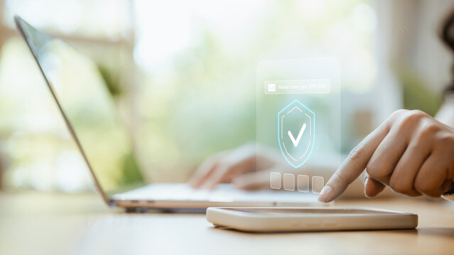 A hand points to a shield interface beside a laptop and smartphone as cybersecurity checks an OTP request and completes verification before granting access to a digital account on the device.
