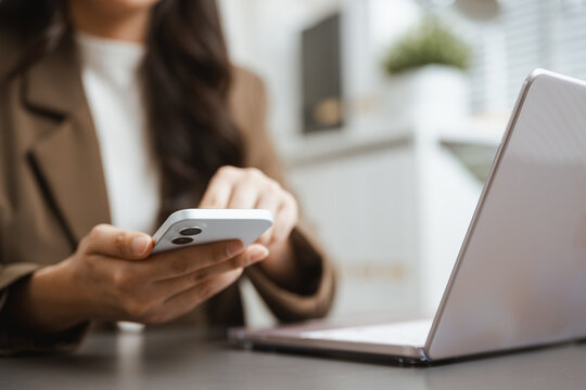 A woman sits beside an open laptop and continues typing on a smartphone while handling mobile communication and routine online tasks from a clean desk in a quiet office during a focused workday.
