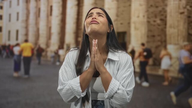 Young woman praying with clasped hands at a historic stone building facade, looking upward among pedestrians in a plaza; plea hope.