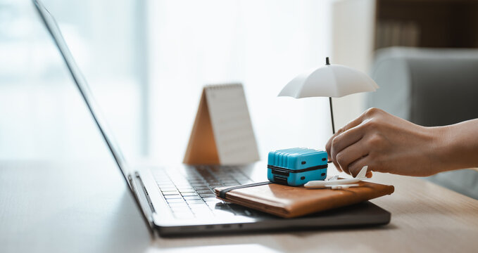 A hand arranges a suitcase and airplane beneath an umbrella beside a laptop to compare travel insurance coverage for medical care and lost luggage before an overseas journey or unexpected delay.
