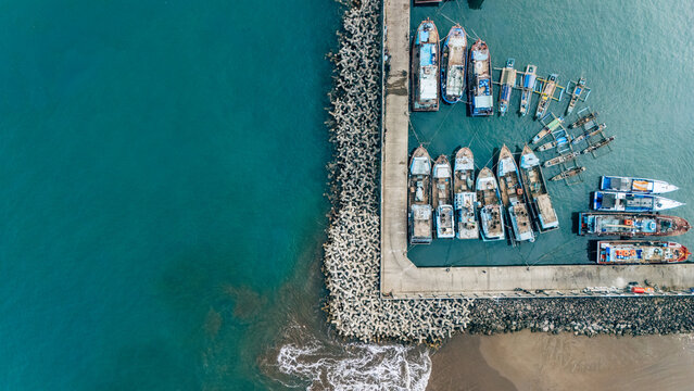 Aerial view of fishing boats docked at a concrete pier with breakwater blocks in the turquoise sea of Palabuhanratu, West Java, Indonesia.