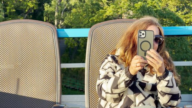 A woman holds up her smartphone while sitting in a deck chair on an open cruise ship deck. She relaxes against a background of lush green trees during a sunny cruise on Lake Ladoga.
