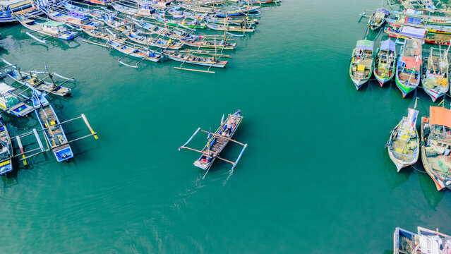 Aerial view of traditional wooden outrigger fishing boats moored in the harbor with one boat navigating the turquoise water in Palabuhanratu, West Java, Indonesia.