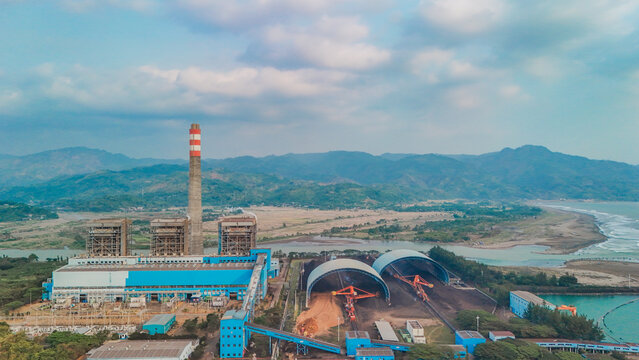 Aerial view of a coal-fired power plant featuring a tall chimney, industrial buildings, and large coal storage domes situated near a river and the coastline