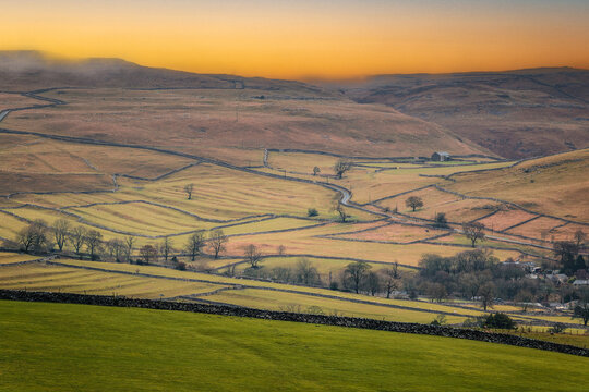 A scenic view of rolling countryside, dry stone walls and winding lanes beneath a golden sunset near Malham, a small village in Yorkshire Dales, UK
