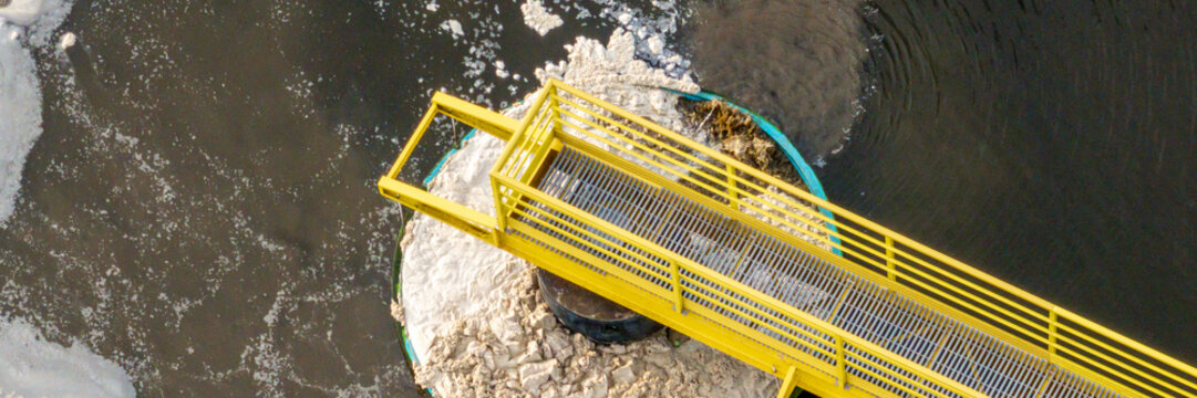 Aerial view of a circular clarifier at a wastewater treatment plant with a yellow metal walkway extending over the water surface in Florianopolis, Santa Catarina, Brazil.