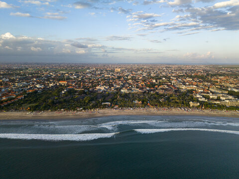 Aerial view of Kuta Beach with waves rolling onto the sandy shore, backed by the dense urban sprawl of Kuta, Bali, Indonesia.