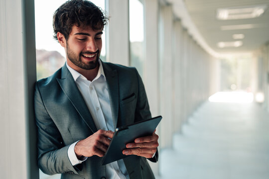 Young businessman smiling, leaning against a wall in a modern office, working with a digital tablet and staying connected