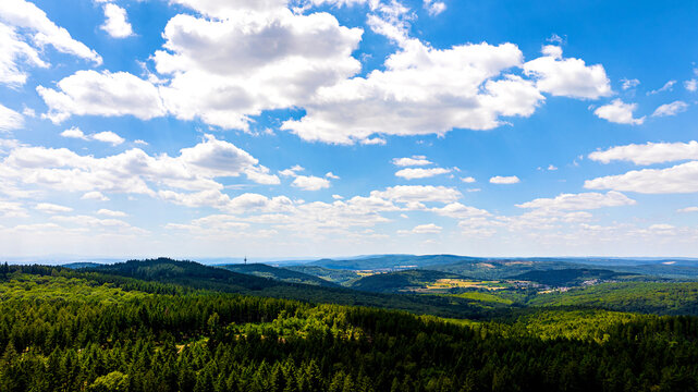 Aerial view of the Grosser Feldberg mountain peak surrounded by dense green forests and rolling hills under a bright blue sky with fluffy white clouds in Schmitten im Taunus, Hessen, Germany.