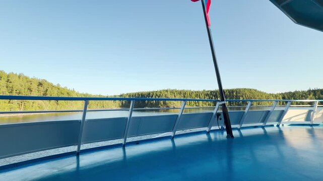 A cruise ship deck moves smoothly past lush pine forests. The scenic view at Valaam on Lake Ladoga evokes a peaceful, serene, and calm feeling under a clear blue sky.