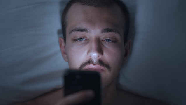 Young Man Lying in Bed at Night Using Smartphone in Dark Room