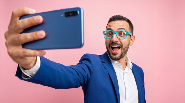 Excited businessman wearing blue suit and colorful glasses taking a smartphone selfie with surprised expression against a bright pink studio background.