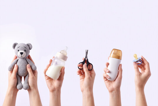 Hands holding baby care essentials including teddy bear, milk bottle, grooming scissors, toiletry container and pacifier against a clean pastel background.