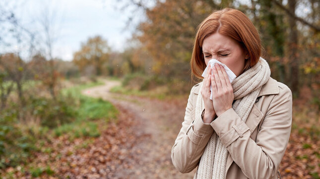 Red haired woman sneezing into a tissue while standing outdoors on an autumn woodland trail with copy space, illustrating seasonal allergy, cold and respiratory health symptoms.