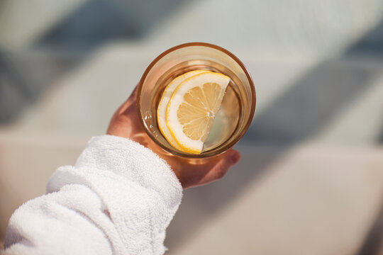 Hand holding a glass of water with a lemon slice, wearing a white towel, sunlight casting shadows on a light surface in a serene setting