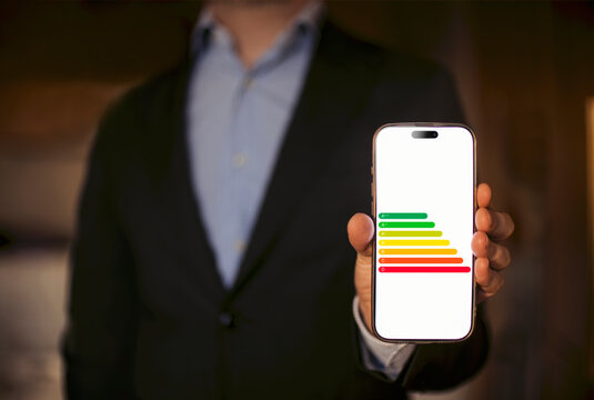 Businessman in a black suit holding smartphone displaying energy efficiency graphic with colored bars representing performance levels in a professional setting