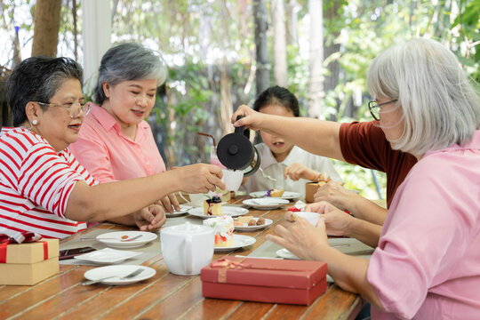 Senior woman pours tea for friend during garden cafe reunion, surrounded by desserts, gift boxes, and granddaughter as shared refreshment strengthens friendship and relaxed social bond, aging society.